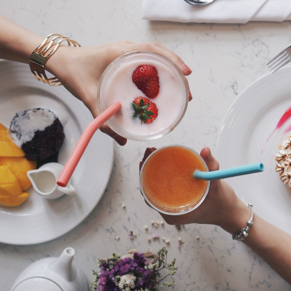 two hands holding two glasses of fruity drinks with ReServe pink and blue silicone reusable drinking straws