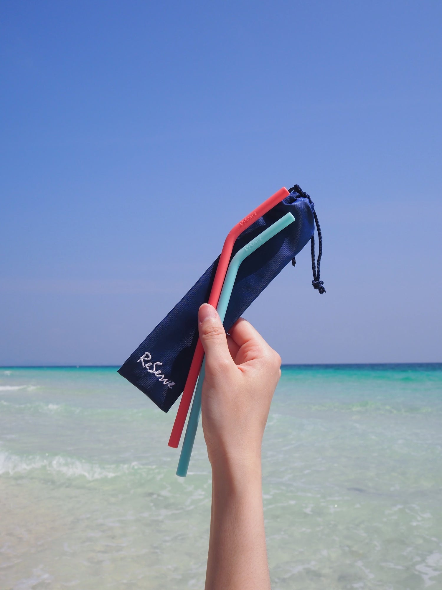 A hand holding a pair of ReServe pink and blue silicone reusable straw with a background of a sea