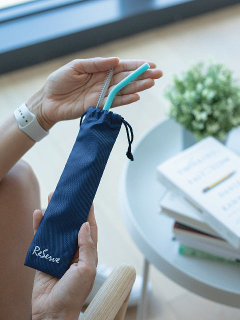 A hand holding the ReServe original starter straw set which includes ReServe blue paradise silicone reusable straw, brush, label, and bag.