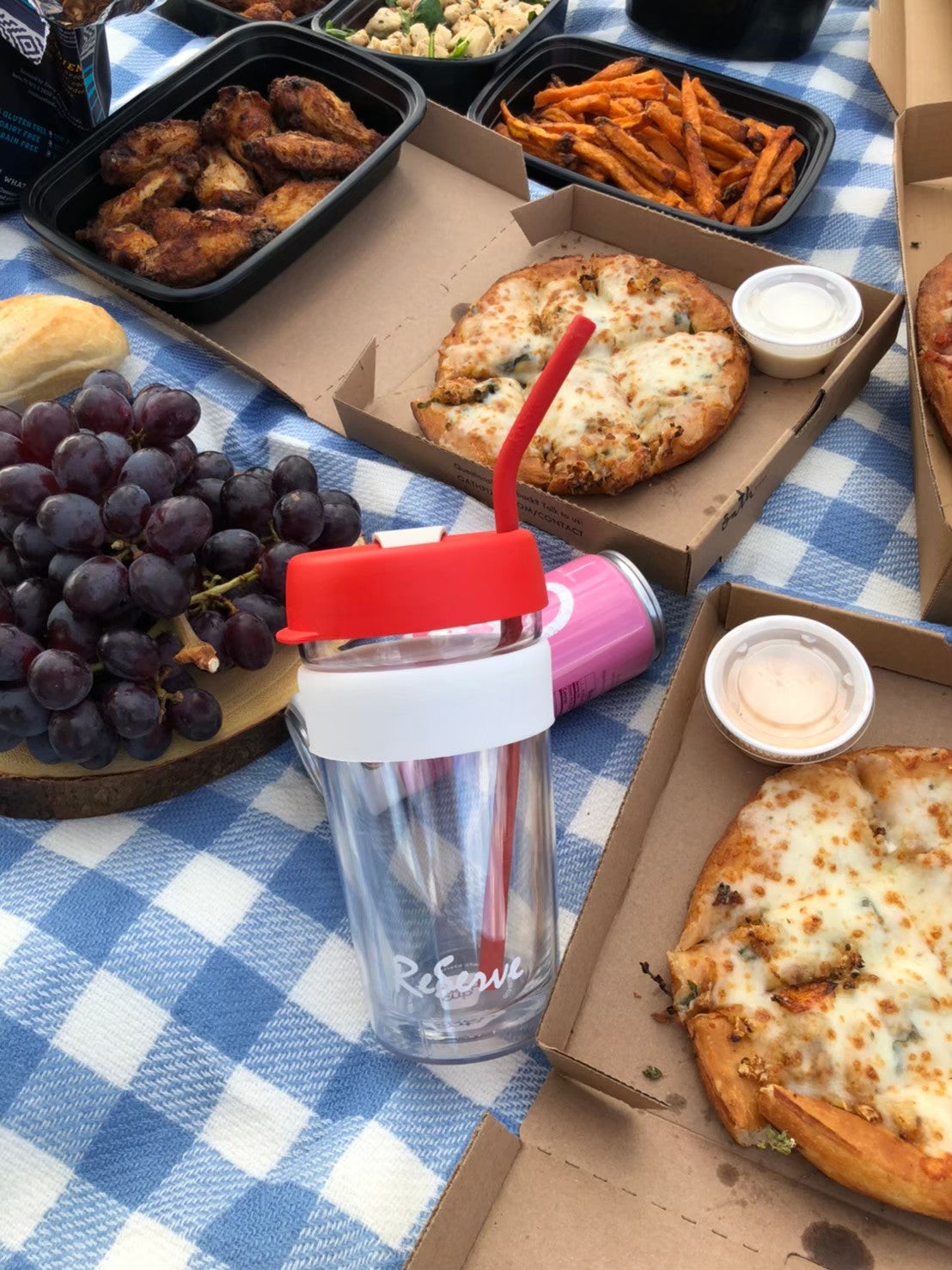 ReServe red reusable cup and red silicone straw on a picnic cloth surrounded by pizza, grape, fries, chicken wings, and other food